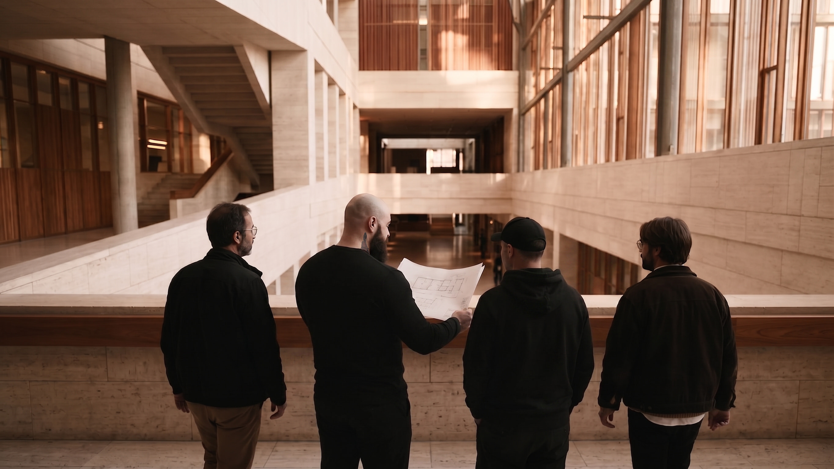 Four people seen from behind at a balustrade, one holding floor plans, looking across a travertine atrium