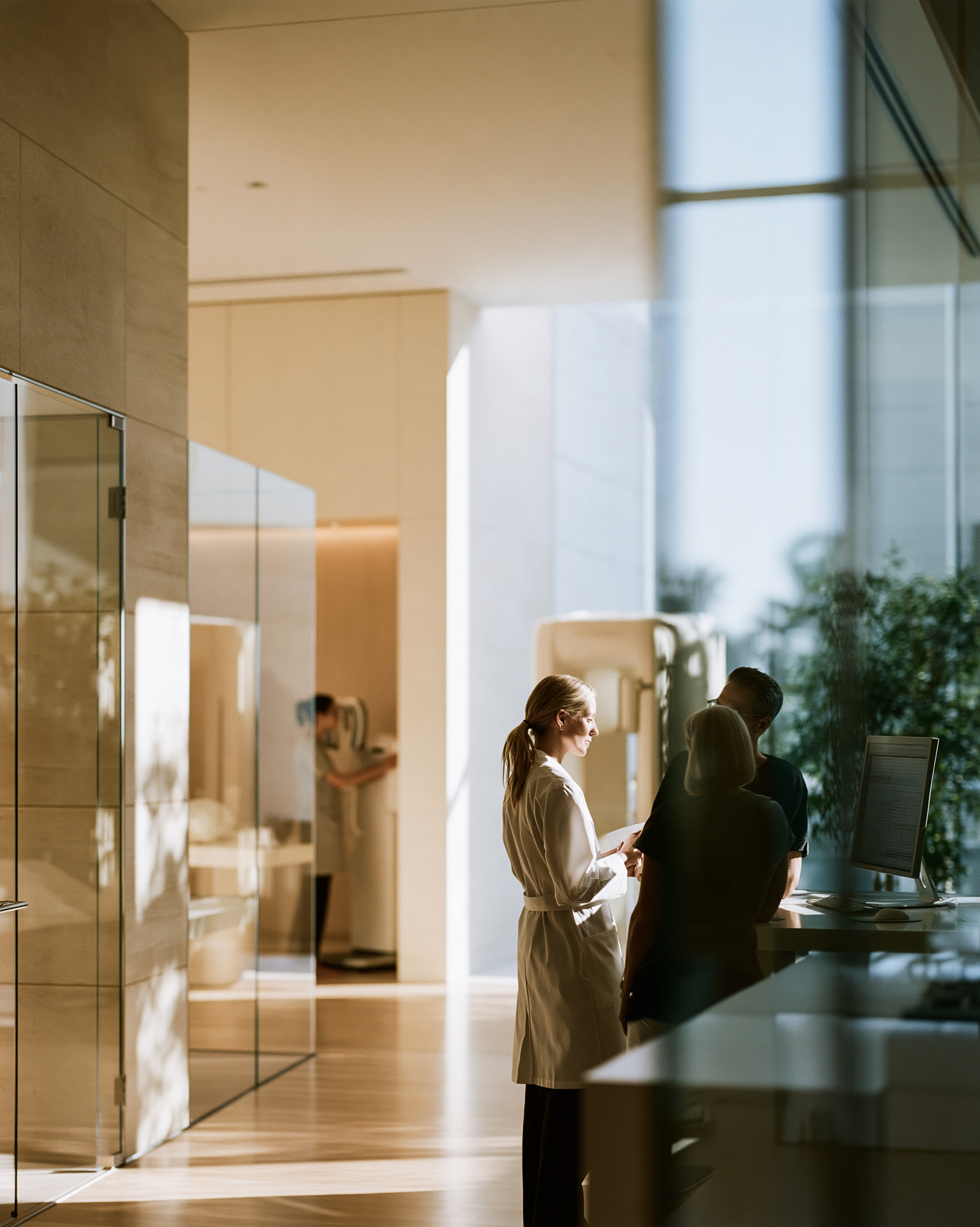 A clinician in a white coat talking with two patients at a reception desk in a sunlit clinic